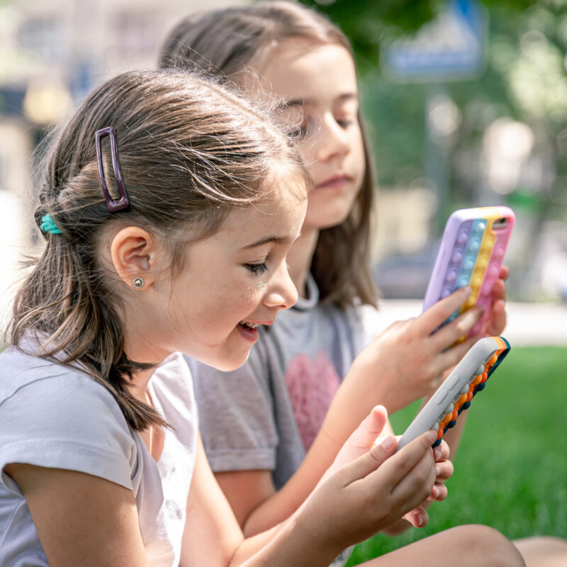 Little funny girls outdoors with phones in a case with pimples pop it, a trendy anti stress toy.